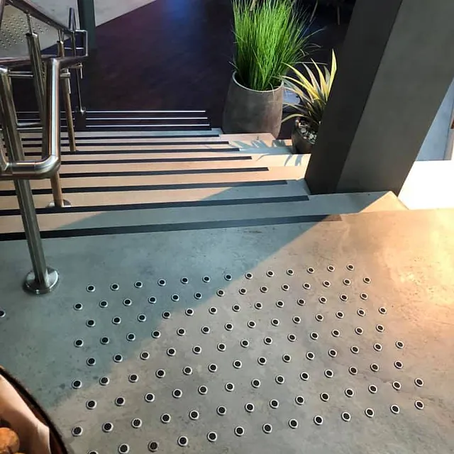 Betongtrapp med innfelte taktile punkter for synshemmede, omgitt av moderne interiør og grønne planter.
