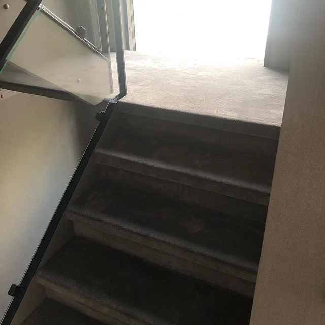 Bratt trapp med mørk vegg-til-vegg-teppe og glassrekkverk, motlys fra dør i toppen av trappen.